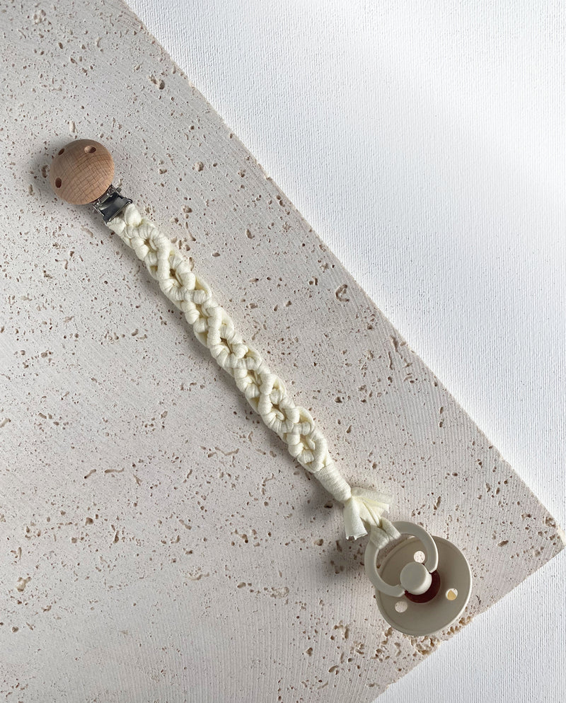 Schnullerband aus Stoff im Makramee Design mit Holzclip in der Farbe creme mit einem Schnuller daran.