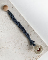 Schnullerband aus Stoff im Makramee Design mit Holzclip in der Farbe dunkelgrau mit einem Schnuller daran.