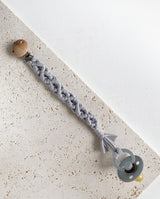 Schnullerband aus Stoff im Makramee Design mit Holzclip in der Farbe hellgrau mit einem grauen Schnuller daran befestigt.