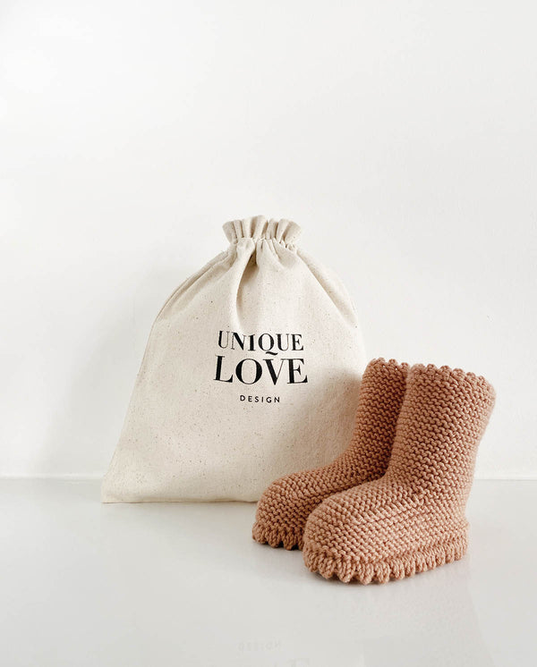 Ein Paar Strickschuhe für Babys mit Häkelkante in der Farbe caramel steht vor einem Leinenbeutel mit Aufschrift "Unique Love Design".