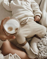 Ein Baby liegt vor der Mama, die einen Kaffee in der Hand hält, und trägt die Strickschuhe in der Farbe creme mit ausgefallener Häkelkante.