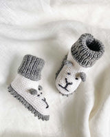 Strickschuhe für Babys in creme mit grauer Häkelkante und Bündchen und mit Löwengesicht-Detail in der Detailansicht.