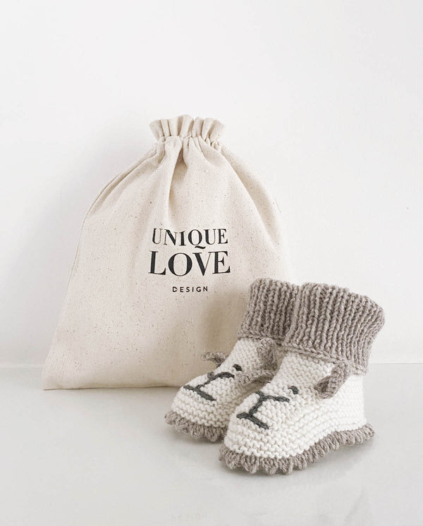 Ein Paar Strickschuhe für Babys in creme mit Häkelkante in der Farbe sand und Bündchen und aufgestickten Löwengesichtern steht vor einem Leinenbeutel mit Aufschrift "Unique Love Design".