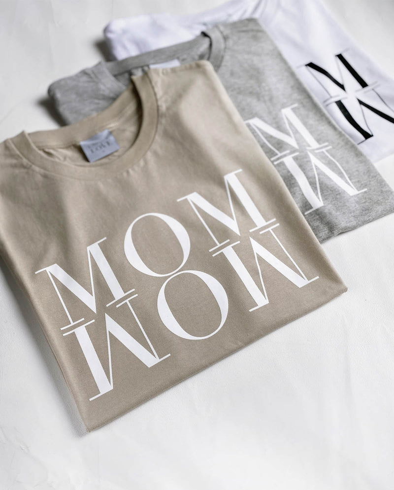Fokus des Schriftzugs MOM WOW auf dem sandfarbenem T-Shirt. Im Hintergrund liegen unter diesem Shirt die beiden weiteren Farbvarianten.