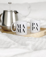 Zwei Keramik Tassen in weiß mit schwarzer Schrift MAMA und PAPA stehen mit einer Teekanne auf einem runden Holztablett.