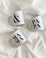 Drei Tassen aus weißem glatten Keramik mit Henkel und schwarzer Aufschrift iegen auf einer weißen Decke. MAMA & PAPA